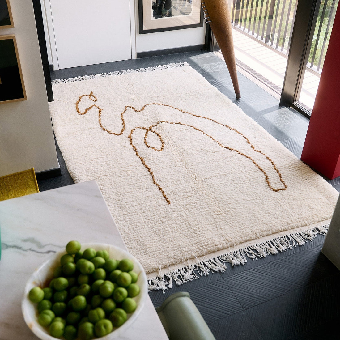 Beige rug with a brown abstract camel design on a gray floor, next to a white table with green fruits.